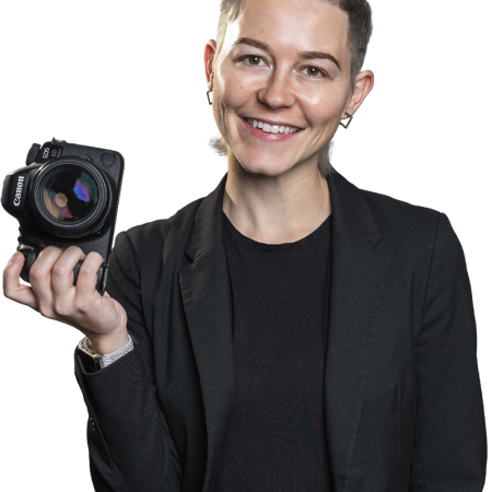 Young androgynous person stands holding a camera in right hand. Smiling and facing the camera wearing all black.