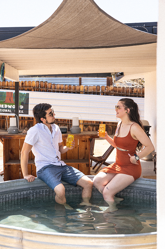 Young woman camera right and young man camera left enjoy beverages while putting their feet in a steel pool outside. There is a beige canopy over head and a wooden bar behind them.