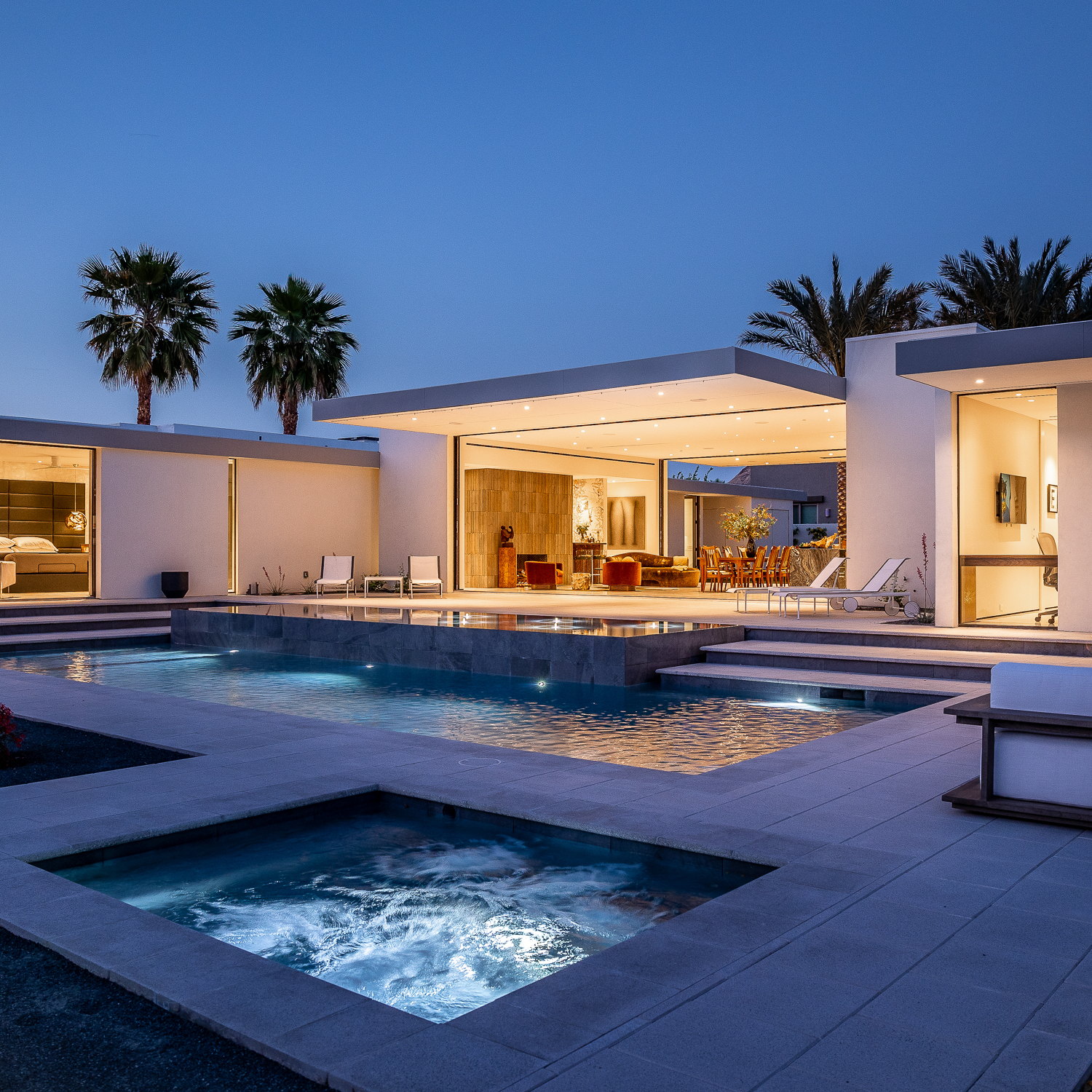 Luxury home photographed at dusk with glowing warm lights inside the home. Pool lights and jacuzzi are on.