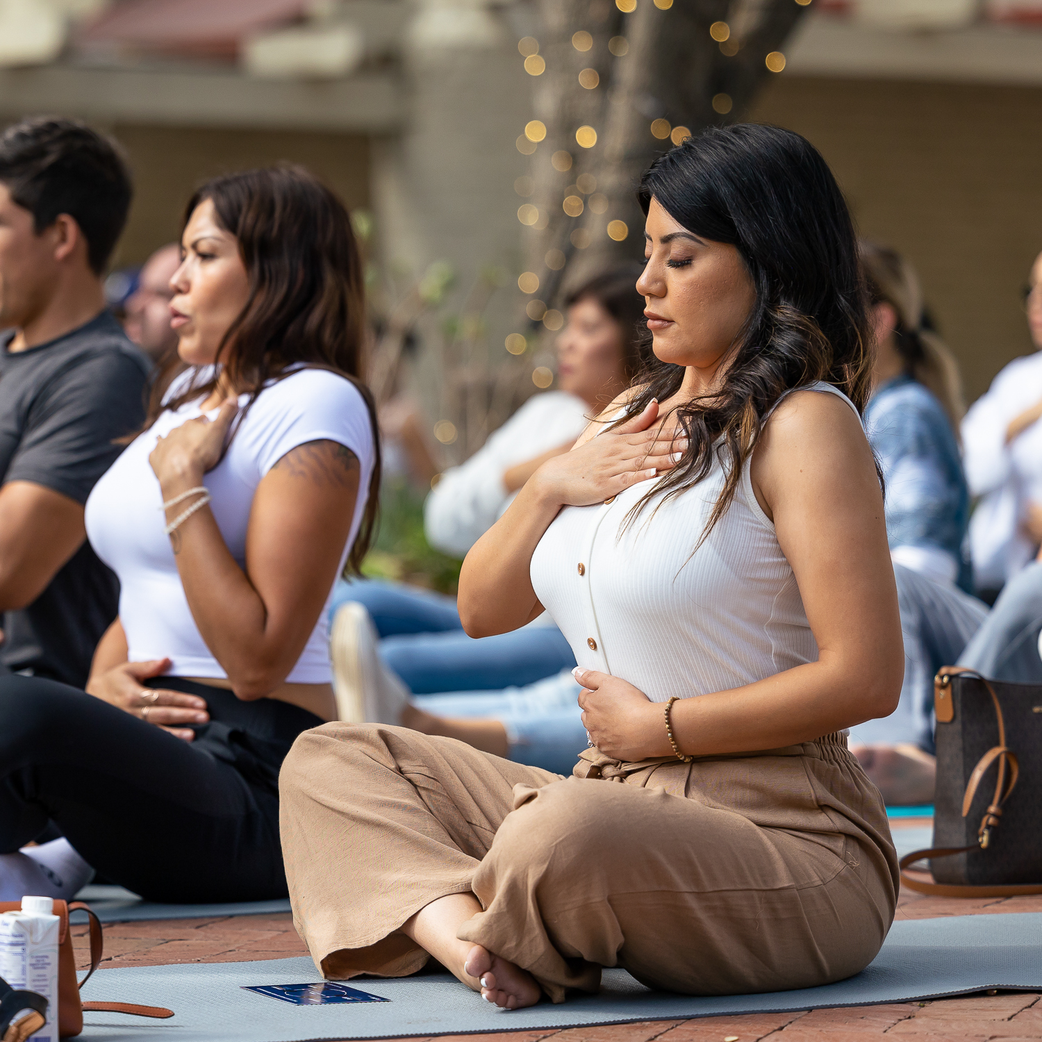 Young woman seated cross legged on a yoga mat among other meditation members. She has one hand over her heart and one over her belly - her eyes are closed.